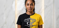 Une femme aux cheveux bouclés se tient contre un mur de marbre, portant le t-shirt Création Clak Cbon de Clak Cbon, divisé en noir et jaune avec le lettrage CLAK CBON et un logo C BON gras sur le devant.