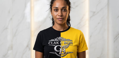 Une femme aux cheveux bouclés se tient contre un mur de marbre, portant le t-shirt Création Clak Cbon de Clak Cbon, divisé en noir et jaune avec le lettrage CLAK CBON et un logo C BON gras sur le devant.